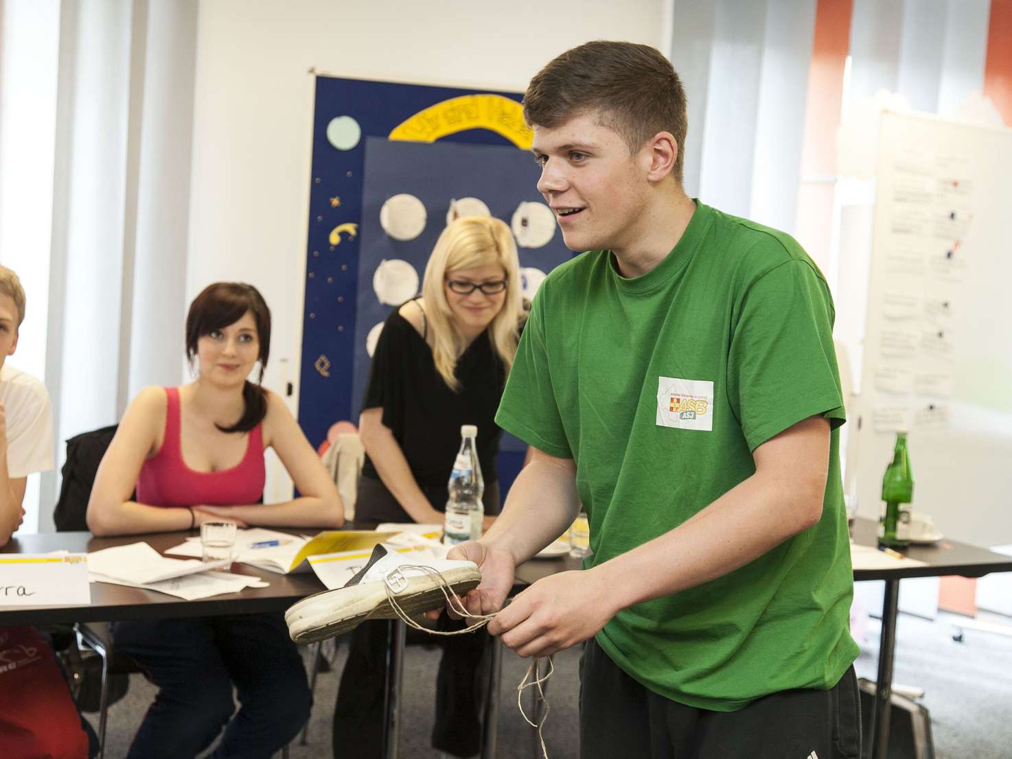 Ein junger Mann im grünen T-Shirt steht in einem Seminarraum und spricht. Im Hintergrund sieht man 3 junge Menschen an einem Tisch sitzen, die ihm zuhören. 