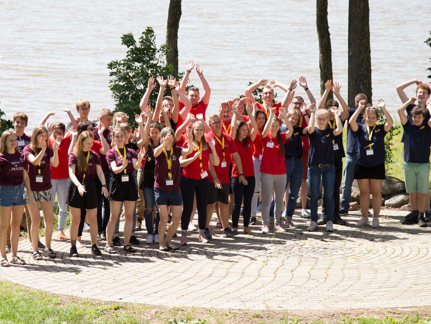 Eine große Gruppe junger Menschen in bunten T-Shirts steht vor einem See. Sie jubeln und heben die Arme in die Luft.